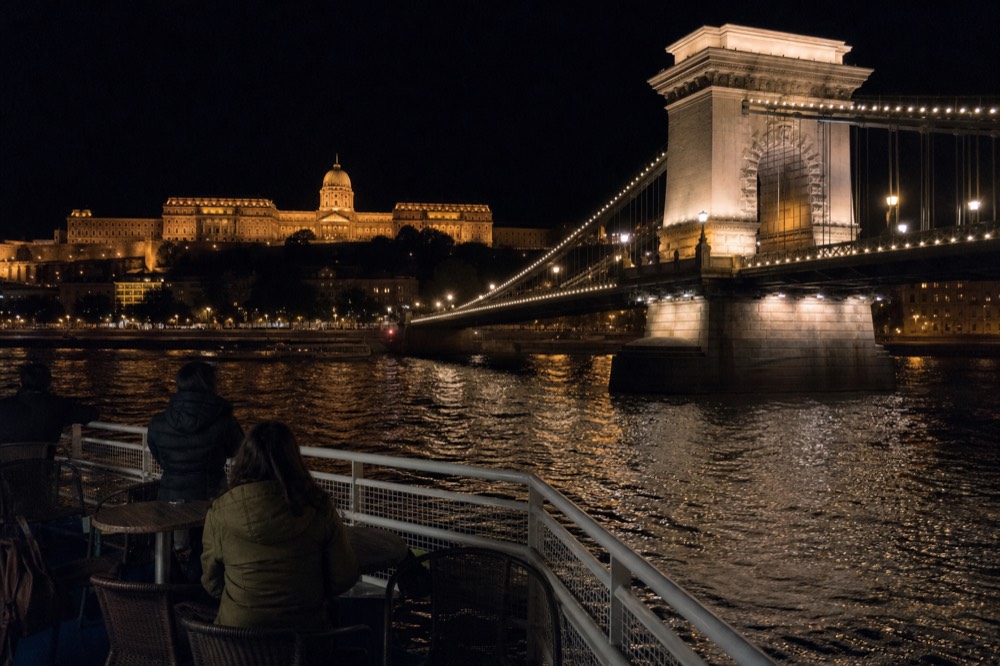 The lit Chain Bridge and Buda Castle seen from a cruise boat on the dark Danube at night, no text, no logos, no people.