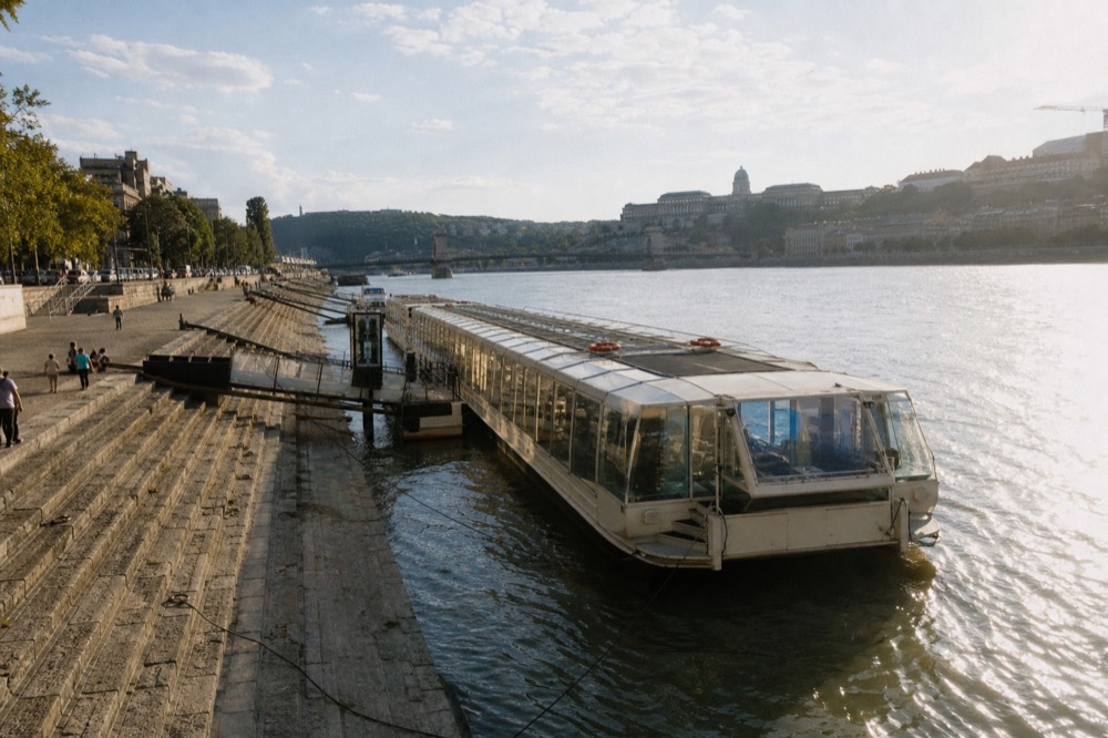 Glass-roofed sightseeing boat moored at a numbered Danube embankment dock in Budapest on a bright afternoon, no text, no logos, no people