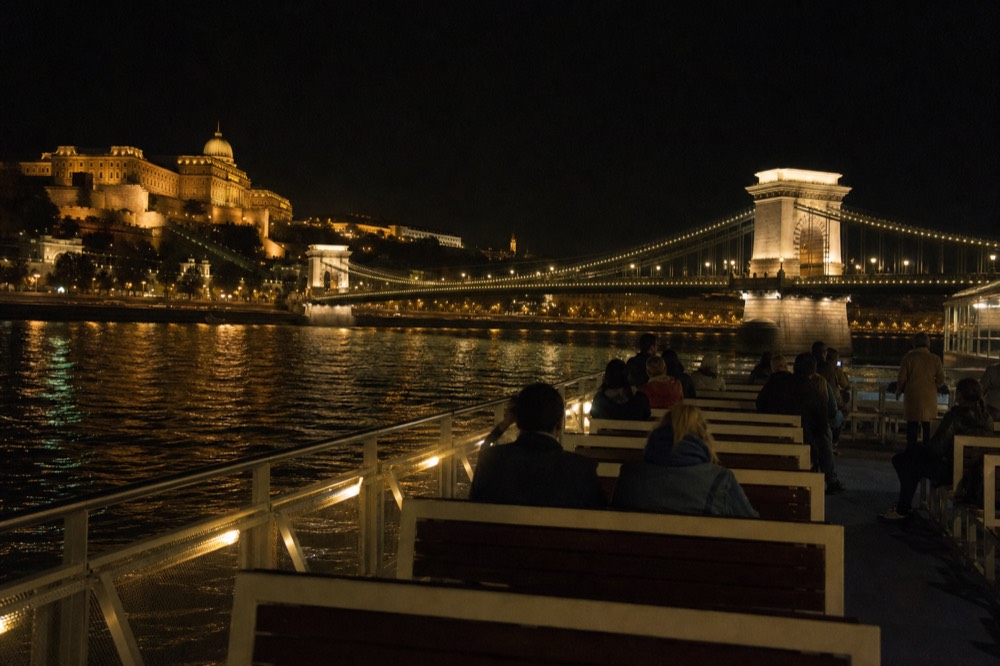 Open top deck of a sightseeing boat gliding along the Danube with the Chain Bridge and Buda Castle lit at night, no text, no logos, no people