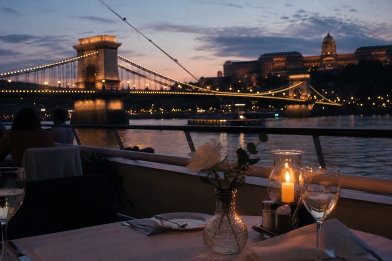 A candlelit table set on the open deck of a Danube dinner-cruise boat at dusk, a lit bridge glowing faintly across the water