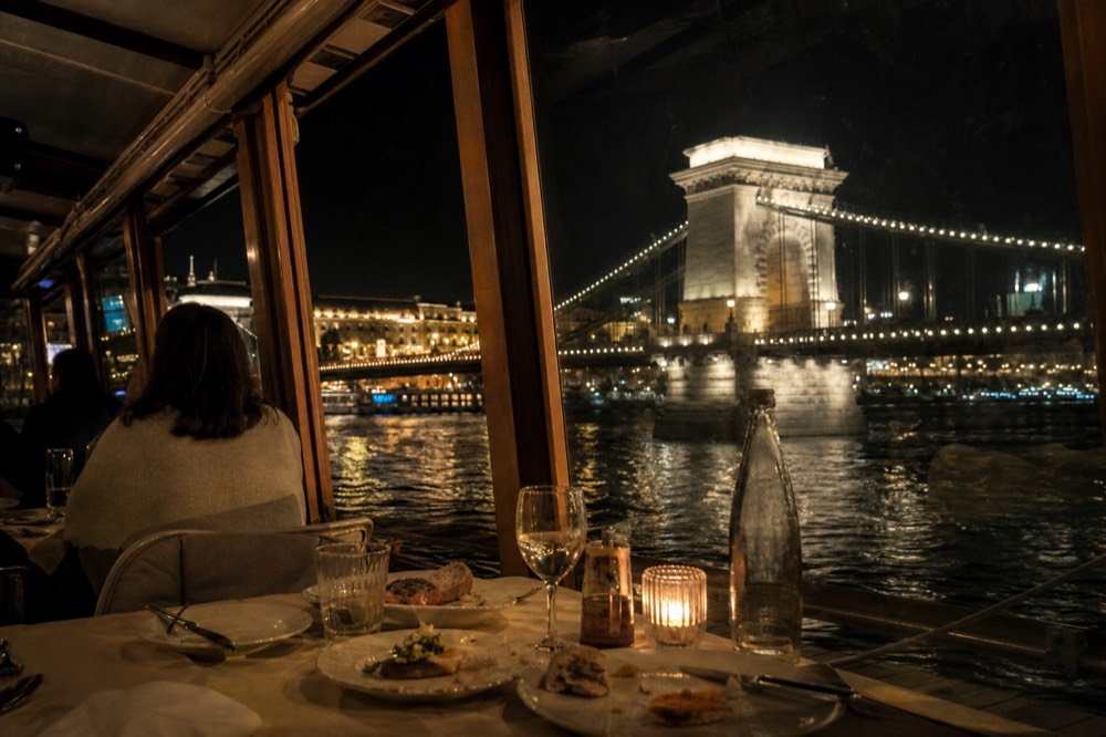 Set dinner table beside a boat window with the Chain Bridge lit beyond, no text, no logos, no people