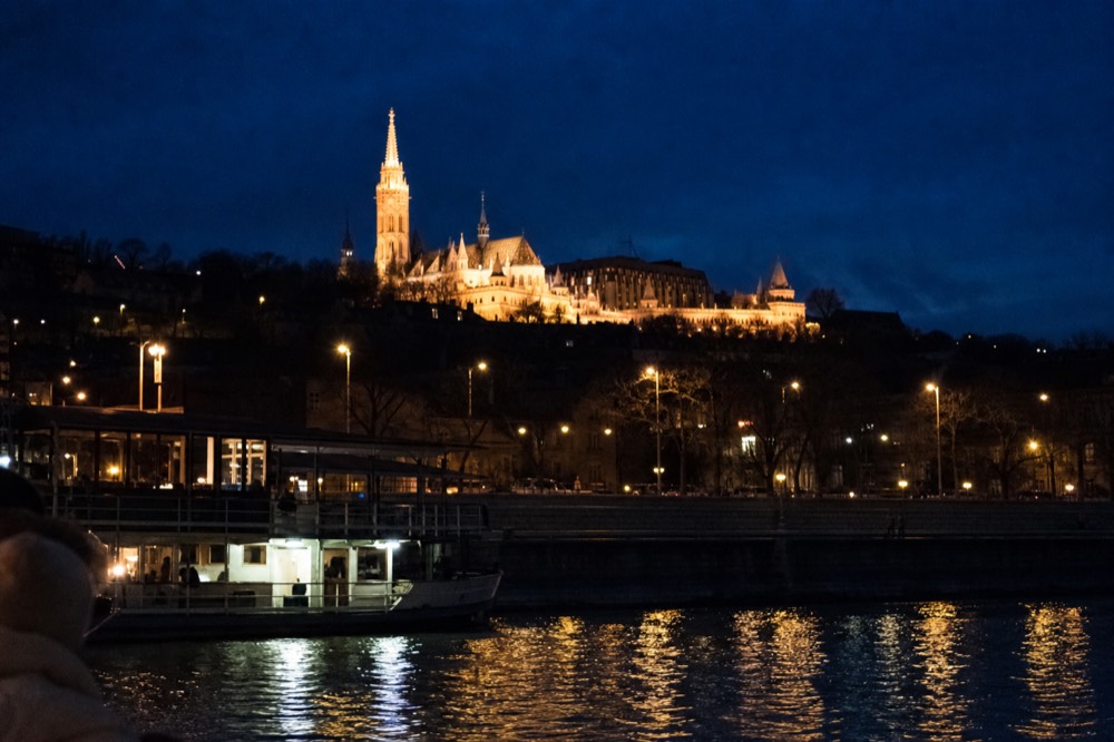 Fisherman's Bastion towers glowing above the Buda hillside viewed from the Danube, no text, no logos, no people