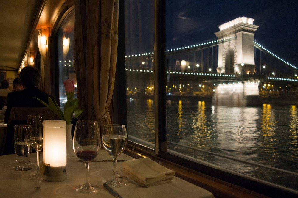 Set dinner table beside a large boat window with the Chain Bridge lit at night beyond the glass, no text, no logos, no people